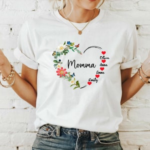 May include: White t-shirt featuring a heart-shaped floral design with the word "Momma" in black script. The heart is adorned with colorful flowers and butterflies. Below the heart are the names Oliva, Anna, Emma, and Emily.
