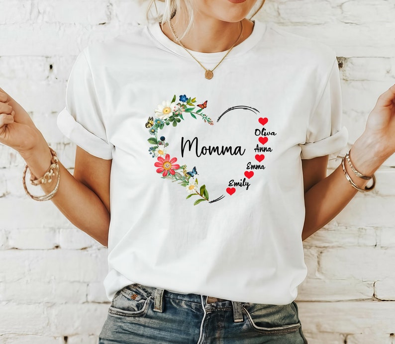 May include: White t-shirt featuring a heart-shaped floral design with the word "Momma" in black script. The heart is adorned with colorful flowers and butterflies. Below the heart are the names Oliva, Anna, Emma, and Emily.