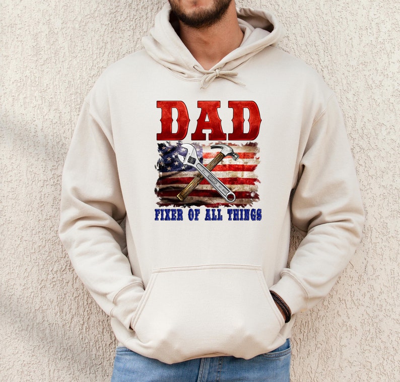 May include: A cream-colored hoodie with a red, white, and blue American flag design. The flag has a wrench and hammer crossed over it. The text "DAD FIXER OF ALL THINGS" is printed below the flag.