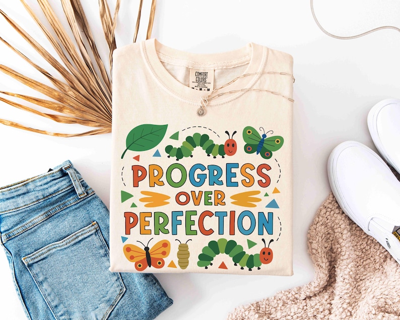 May include: Cream-colored t-shirt with the words "PROGRESS OVER PERFECTION" in colorful letters. The shirt features cartoon caterpillars, butterflies, and a leaf design. The shirt is folded and placed on a white surface with blue jeans and white shoes.