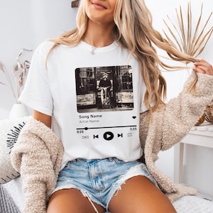 May include: White t-shirt with a black and white image of a person sitting on a bench. The image is framed with a black border and has a music player interface below it with the text "Song Name" and "Artist Name".