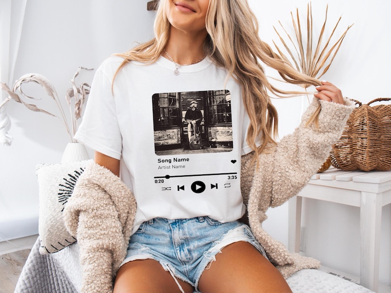 May include: White t-shirt with a black and white image of a person sitting on a bench. The image is framed with a black border and has a music player interface below it with the text "Song Name" and "Artist Name".