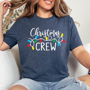 Puede incluir: Una camiseta azul marino con las palabras "Christmas CREW" en escritura blanca y una cadena de luces navide&ntilde;as de colores. La camiseta es de cuello redondo y manga corta.