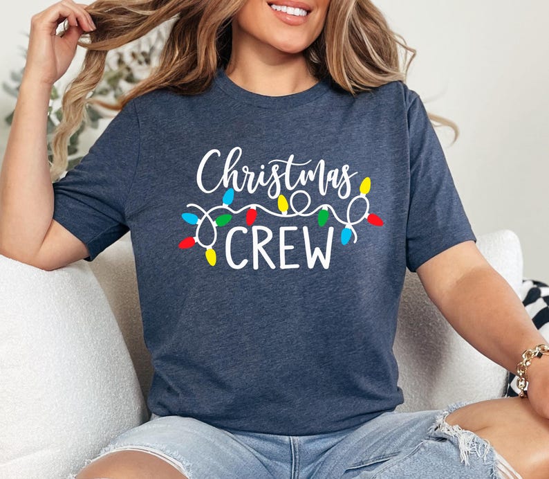 Puede incluir: Una camiseta azul marino con las palabras "Christmas CREW" en escritura blanca y una cadena de luces navide&ntilde;as de colores. La camiseta es de cuello redondo y manga corta.