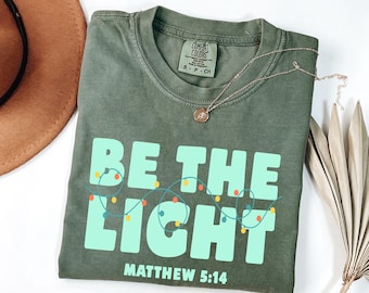 May include: Olive green t-shirt with the text "BE THE LIGHT" in light blue, with a string of colorful lights. The text "Matthew 5:14" is below the main text. A gold necklace with a pendant is on the shirt.