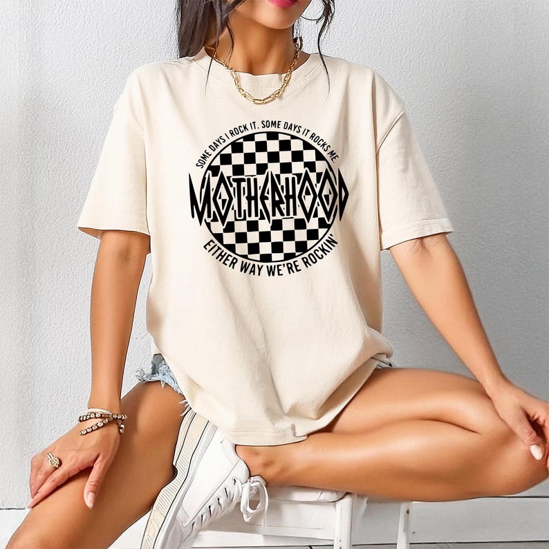 May include: A white t-shirt with a black and white checkered circle graphic. The graphic has the text "SOME DAYS I ROCK IT. SOME DAYS IT ROCKS ME. MOTHERHOOD EITHER WAY WE'RE ROCKIN'" around the checkered circle.
