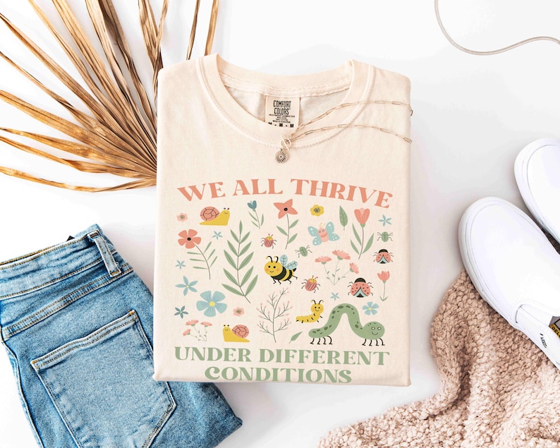 May include: Cream-colored t-shirt with the text "WE ALL THRIVE UNDER DIFFERENT CONDITIONS" and a colorful graphic of flowers, insects, and a caterpillar. The shirt is folded next to blue jeans, white sneakers, and a beige blanket.