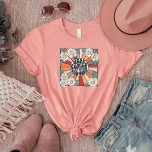 May include: A coral-colored t-shirt with a retro graphic that reads "Here Comes the Sun." The design features a sunburst with orange, brown, and cream rays, surrounded by white daisies and green leaves. The shirt is folded.