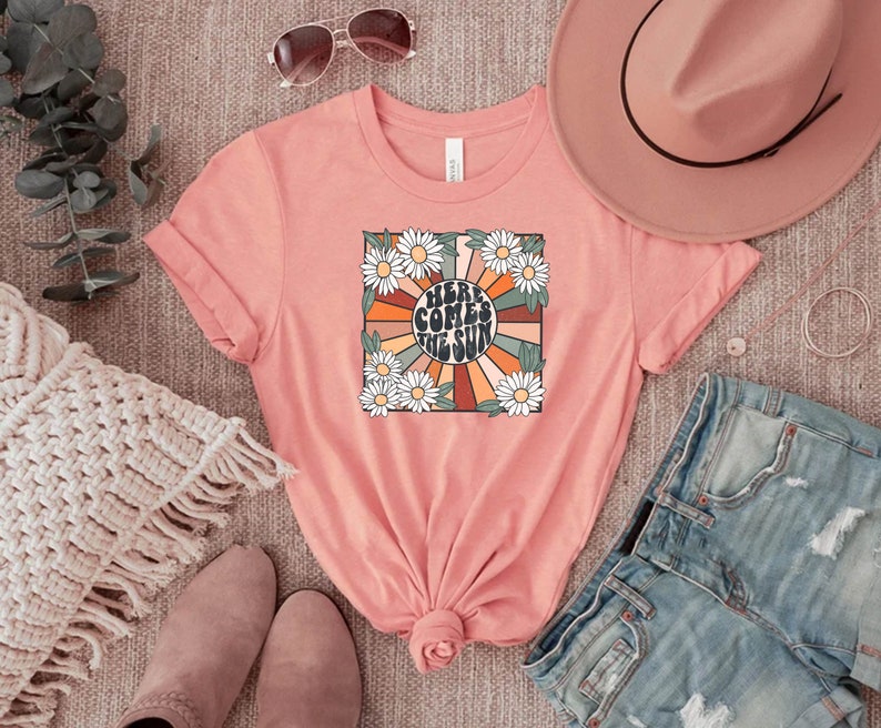May include: A coral-colored t-shirt with a retro graphic that reads "Here Comes the Sun." The design features a sunburst with orange, brown, and cream rays, surrounded by white daisies and green leaves. The shirt is folded.