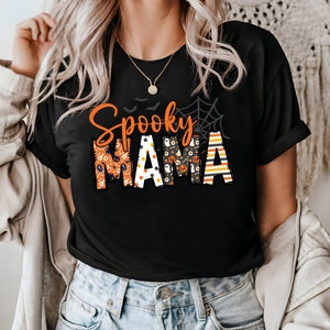 以下が含まれることがあります： オレンジ色と模様の文字で「Spooky Mama」と書かれた黒いTシャツ。デザインには、コウモリ、クモの巣、ハロウィーンをテーマにした模様が含まれています。金のネックレスが見えます。