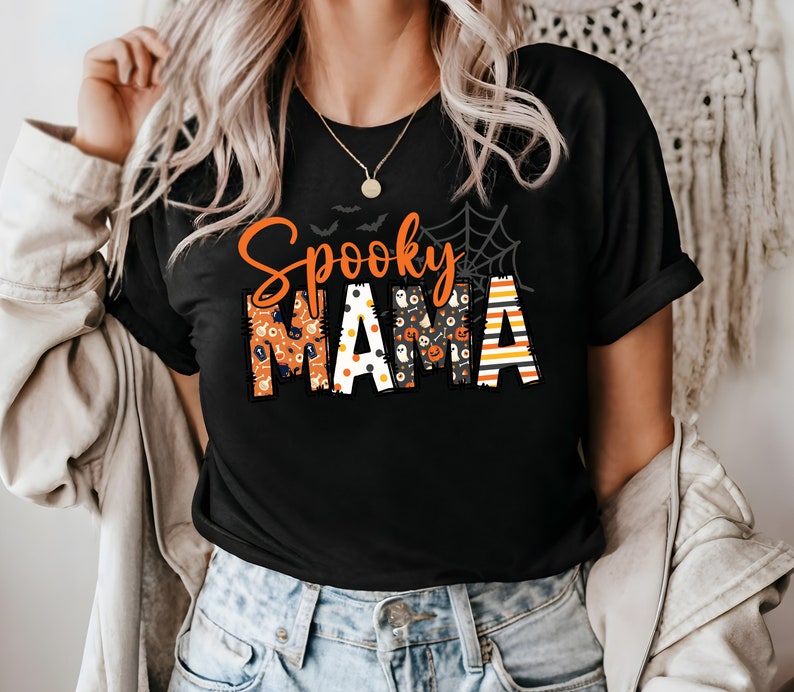 以下が含まれることがあります： オレンジ色と模様の文字で「Spooky Mama」と書かれた黒いTシャツ。デザインには、コウモリ、クモの巣、ハロウィーンをテーマにした模様が含まれています。金のネックレスが見えます。