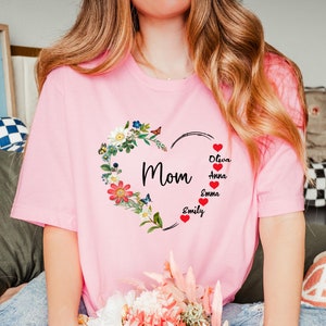May include: Pink t-shirt featuring a floral heart design with the word "Mom" in the center. The heart is adorned with flowers, butterflies, and the names Olivia, Anna, Emma, and Emily. A personalized gift.