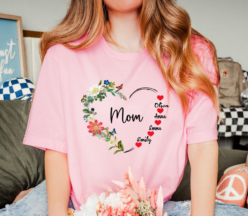 May include: Pink t-shirt featuring a floral heart design with the word "Mom" in the center. The heart is adorned with flowers, butterflies, and the names Olivia, Anna, Emma, and Emily. A personalized gift.