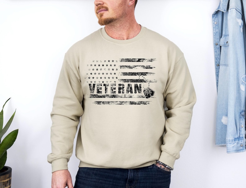 Peut inclure: Un sweat-shirt beige avec un motif de drapeau am&eacute;ricain vieilli et le mot "Veteran" imprim&eacute; en noir. Des m&eacute;dailles militaires pendent en bas du drapeau.