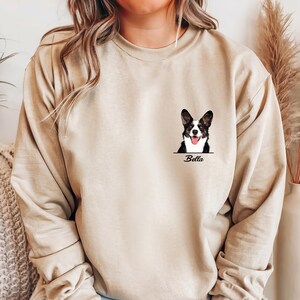 May include: Beige long-sleeve sweatshirt featuring a black and white dog portrait with the name "Bella" below. The dog has its tongue out. The sweatshirt is a neutral color and has a relaxed fit.