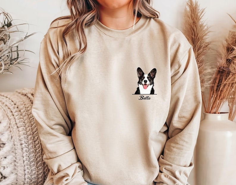 May include: Beige long-sleeve sweatshirt featuring a black and white dog portrait with the name "Bella" below. The dog has its tongue out. The sweatshirt is a neutral color and has a relaxed fit.
