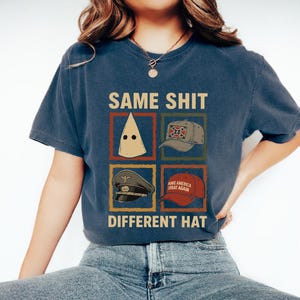 May include: Navy blue t-shirt with the text "SAME SHIT DIFFERENT HAT" and images of different hats. The hats include a white hood, a Confederate flag baseball cap, a military hat, and a red "MAKE AMERICA GREAT AGAIN" hat.