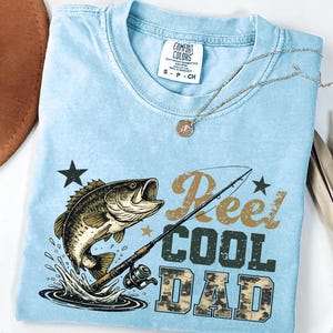 May include: Light blue Comfort Colors t-shirt with a graphic of a leaping fish, fishing rod, and the text "Reel Cool Dad." A silver necklace with a pendant rests on the shirt. The shirt is size small.