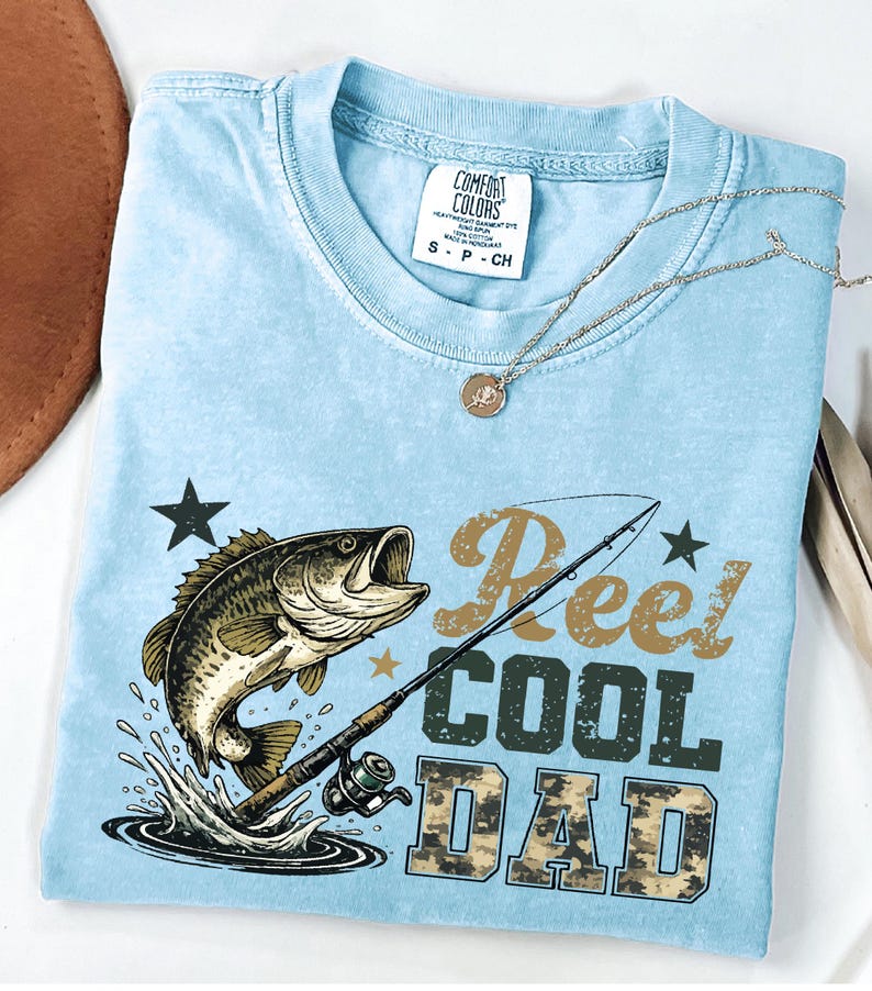 May include: Light blue Comfort Colors t-shirt with a graphic of a leaping fish, fishing rod, and the text "Reel Cool Dad." A silver necklace with a pendant rests on the shirt. The shirt is size small.