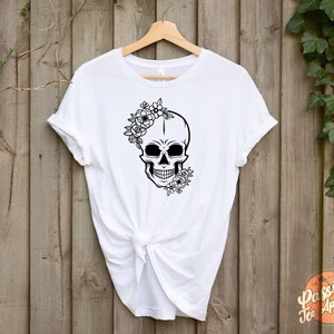 May include: White t-shirt featuring a black skull design adorned with floral accents. The skull has a floral crown and additional flowers on the side. The shirt is hanging on a wooden background.