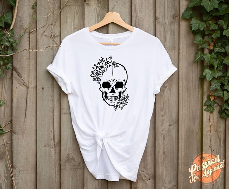 May include: White t-shirt featuring a black skull design adorned with floral accents. The skull has a floral crown and additional flowers on the side. The shirt is hanging on a wooden background.