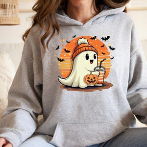 May include: A grey hoodie featuring a cute cartoon ghost wearing a hat and holding a pumpkin and a drink. The ghost is in front of a sunset with bats flying around it. The text on the hoodie says "Boo!"