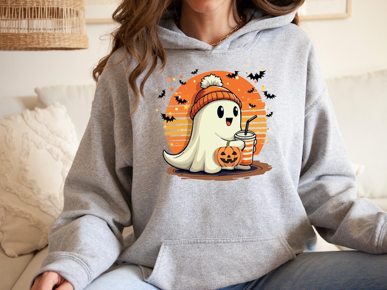 May include: A grey hoodie featuring a cute cartoon ghost wearing a hat and holding a pumpkin and a drink. The ghost is in front of a sunset with bats flying around it. The text on the hoodie says "Boo!"