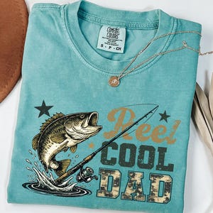 May include: A turquoise Comfort Colors t-shirt with a graphic of a leaping fish, fishing rod, and the words "Reel Cool Dad." The shirt has a crew neck and short sleeves. A gold necklace with a pendant is draped across the shirt.