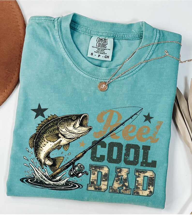 May include: A turquoise Comfort Colors t-shirt with a graphic of a leaping fish, fishing rod, and the words "Reel Cool Dad." The shirt has a crew neck and short sleeves. A gold necklace with a pendant is draped across the shirt.