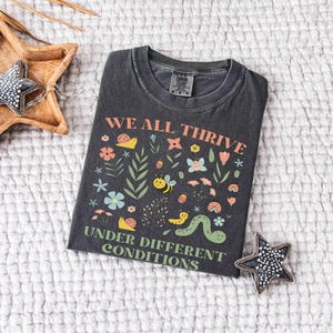 May include: Dark gray t-shirt with the text "WE ALL THRIVE UNDER DIFFERENT CONDITIONS" and a colorful graphic of flowers, bees, and a caterpillar. The shirt is laid on a white textured surface, with a wooden star-shaped tray and a beaded star-shaped decoration.