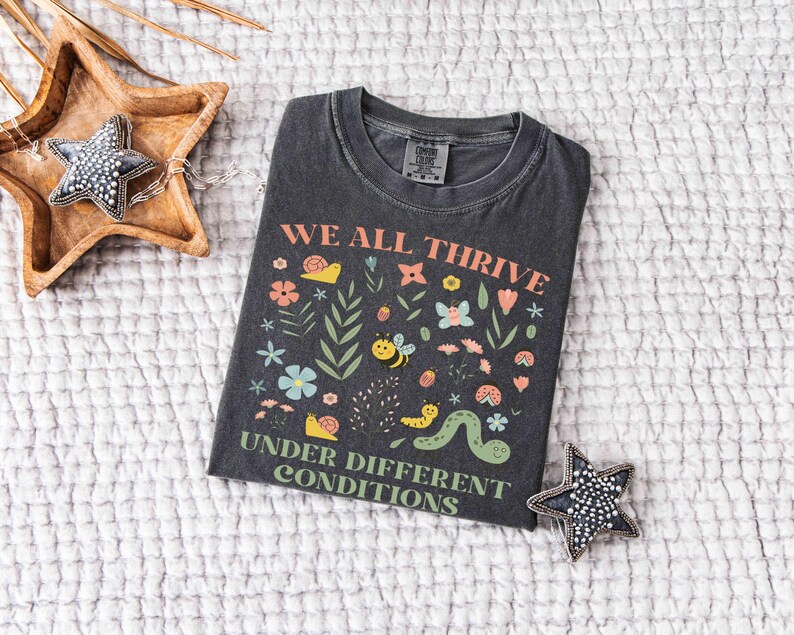 May include: Dark gray t-shirt with the text "WE ALL THRIVE UNDER DIFFERENT CONDITIONS" and a colorful graphic of flowers, bees, and a caterpillar. The shirt is laid on a white textured surface, with a wooden star-shaped tray and a beaded star-shaped decoration.