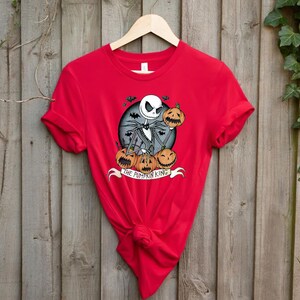 May include: A red t-shirt featuring a graphic of Jack Skellington from The Nightmare Before Christmas. The design includes Jack holding a pumpkin, surrounded by other pumpkins, with the text "THE PUMPKIN KING" on a banner.