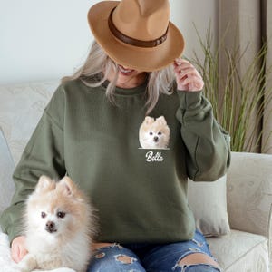 May include: Olive green sweatshirt featuring a Pomeranian dog portrait with the name "Bella" printed below. The dog's image is in a pocket-like design. The sweatshirt is worn by a person wearing a brown hat.