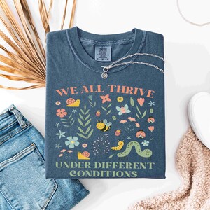 May include: A blue t-shirt with the words "WE ALL THRIVE UNDER DIFFERENT CONDITIONS" and a colorful graphic of flowers, insects, and plants. The shirt is paired with blue jeans, white shoes, and a silver necklace.