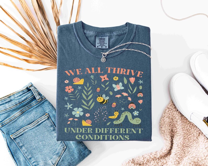 May include: A blue t-shirt with the words "WE ALL THRIVE UNDER DIFFERENT CONDITIONS" and a colorful graphic of flowers, insects, and plants. The shirt is paired with blue jeans, white shoes, and a silver necklace.