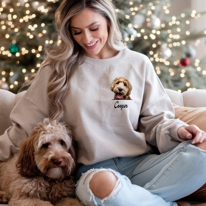 May include: Beige sweatshirt featuring a dog portrait and the name "Cooper" printed below. The dog has brown and tan fur. The sweatshirt is worn by a person. A dog with similar fur is in the foreground.