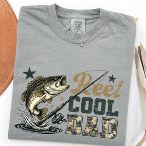 May include: A gray t-shirt with a fishing theme. The shirt features a graphic of a fish jumping out of water, a fishing rod, and the text "Reel Cool Dad" in a mix of fonts and colors. A gold necklace with a star pendant is on the shirt.