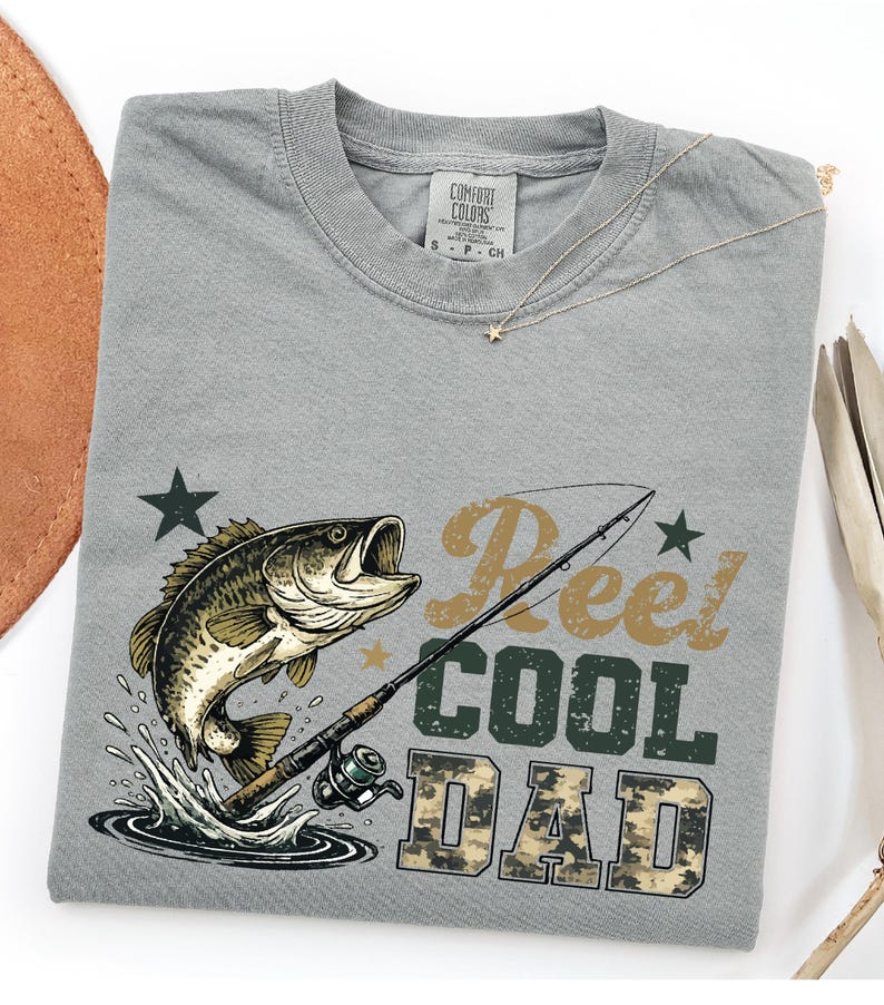 May include: A gray t-shirt with a fishing theme. The shirt features a graphic of a fish jumping out of water, a fishing rod, and the text "Reel Cool Dad" in a mix of fonts and colors. A gold necklace with a star pendant is on the shirt.