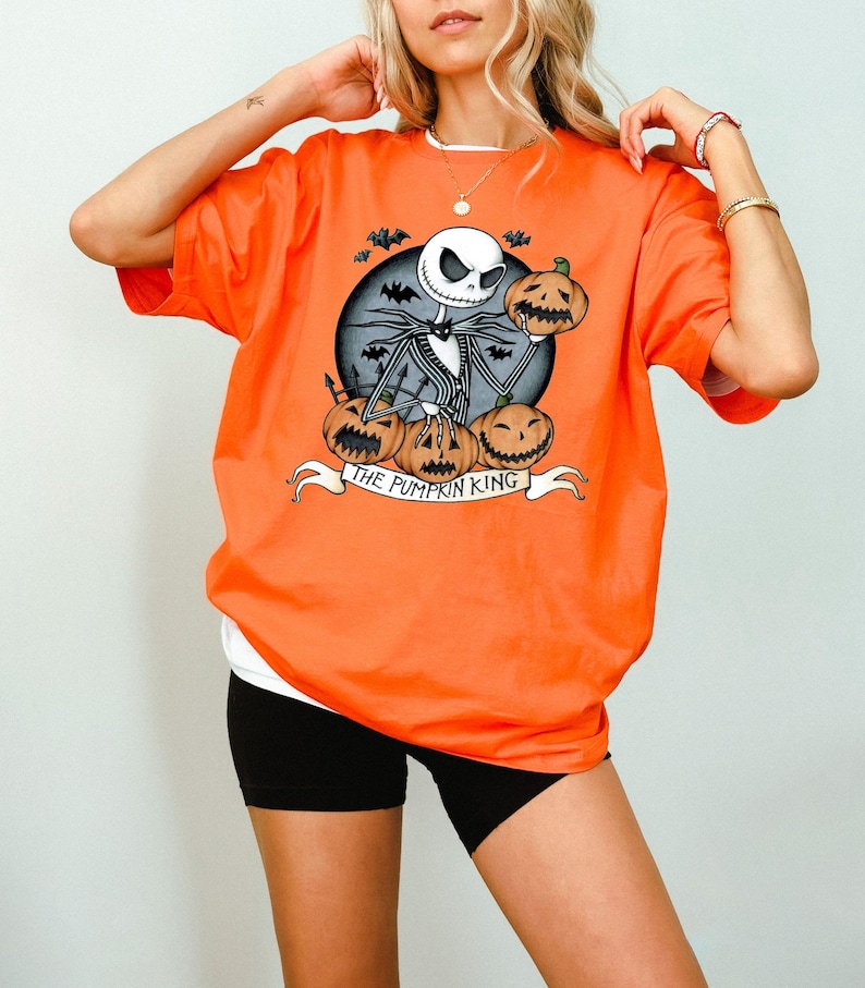 May include: Orange short-sleeved t-shirt featuring a graphic of Jack Skellington from The Nightmare Before Christmas, holding a pumpkin. The design includes bats, pumpkins, and the text "THE PUMPKIN KING" on a banner.