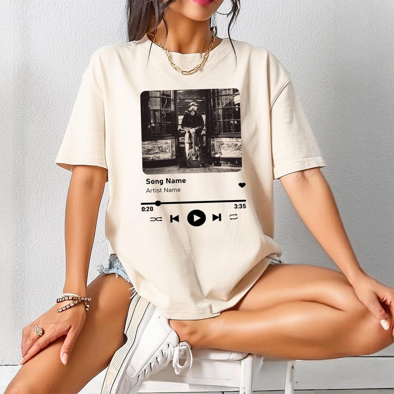 May include: A white t-shirt with a black and white image of a person playing a guitar. The image is framed with a black border and has a play button, rewind button, and fast forward button below it. The text "Song Name" and "Artist Name" are below the image. The text "0:20" and "3:35" are at the bottom left and right corners of the image.