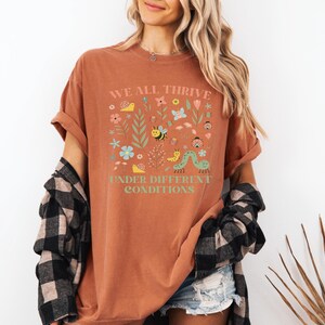 May include: Rust-colored t-shirt with the text "WE ALL THRIVE UNDER DIFFERENT CONDITIONS" and a colorful floral and insect design. The shirt is paired with denim shorts and a black and white plaid shirt.