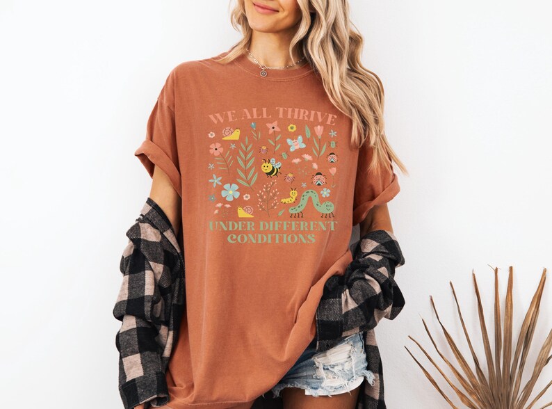 May include: Rust-colored t-shirt with the text "WE ALL THRIVE UNDER DIFFERENT CONDITIONS" and a colorful floral and insect design. The shirt is paired with denim shorts and a black and white plaid shirt.
