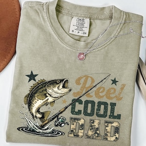 May include: Olive green t-shirt with a graphic of a fish jumping out of water, a fishing rod, and the text "Reel Cool Dad." The shirt has a crew neck and short sleeves. A silver necklace with a pink pendant is on top of the shirt.