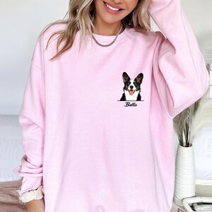 May include: A light pink sweatshirt featuring a corgi dog graphic with the name "Bella" printed below. The corgi is black and white with its tongue out. The sweatshirt has a crew neck and long sleeves.