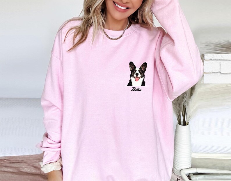 May include: A light pink sweatshirt featuring a corgi dog graphic with the name "Bella" printed below. The corgi is black and white with its tongue out. The sweatshirt has a crew neck and long sleeves.