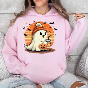 May include: A pink hoodie featuring a cute cartoon ghost wearing a pumpkin hat and holding a pumpkin and a drink. The ghost is in front of a sunset with bats flying around it.