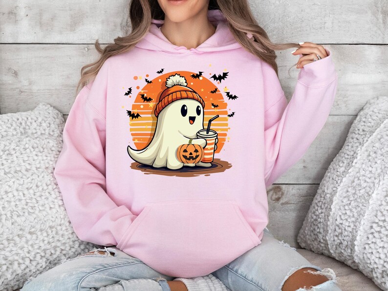 May include: A pink hoodie featuring a cute cartoon ghost wearing a pumpkin hat and holding a pumpkin and a drink. The ghost is in front of a sunset with bats flying around it.