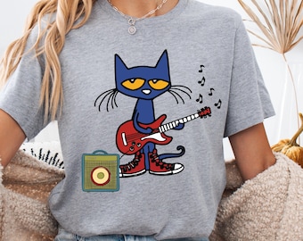 May include: A heather gray short-sleeve t-shirt featuring a graphic of a blue cartoon cat playing a red electric guitar while wearing red sneakers. A small yellow and blue guitar amplifier sits next to the cat, with black musical notes floating nearby.