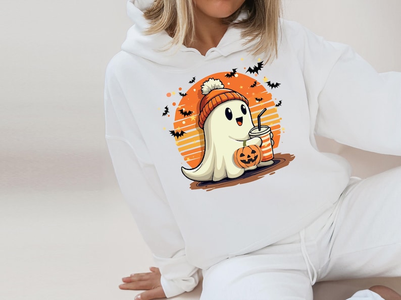 May include: White hoodie featuring a cute cartoon ghost wearing a hat and holding a pumpkin and a drink. The ghost is in front of an orange and yellow sunset with bats flying around it.
