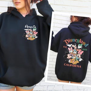 May include: Black hooded sweatshirt with a graphic of Mickey Mouse and friends. The text "Amelia" is printed on the front of the sweatshirt. The back of the sweatshirt has a graphic of Mickey Mouse and friends with the text "Disneyland Est. 1955 California".